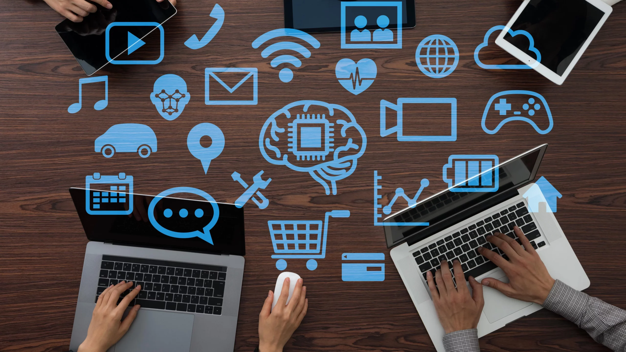 Top-down view of a wooden table with multiple hands using laptops and tablets, surrounded by floating blue digital icons representing technology, communication, cloud computing, AI, gaming, shopping, and data analytics